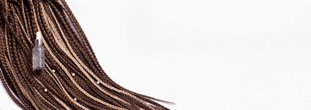 Banner. Thin brown African braids and a jar of care oil on a white background. Hair close-up, braided dreadlocks, afro style
