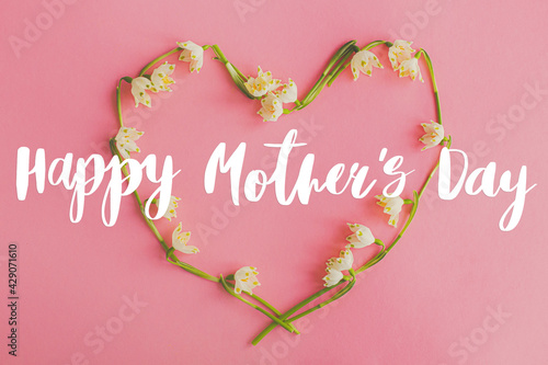 Happy mother's day greeting card. Happy mother's day text and floral heart flat lay on pink paper. Stylish floral greetings. Handwritten lettering on heart of spring flowers on pink. Mothers day
