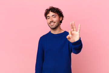 hispanic handsome man smiling and looking friendly, showing number three or third with hand...