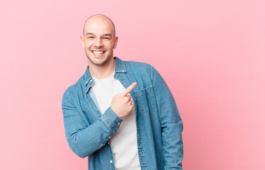 bald man smiling cheerfully, feeling happy and pointing to the side and upwards, showing object in...