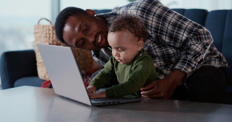 Afro-american bonding family with baby sitting in living room. Young african dad teaching little child to use computer videochatting on laptop together.