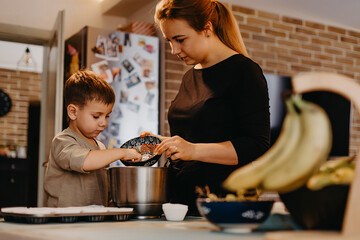 Mom and her son helping to prepare cupcakes in the kitchen. The boy with plaster on his hand mixes the dough and puts it in baking tins, and mom tells
