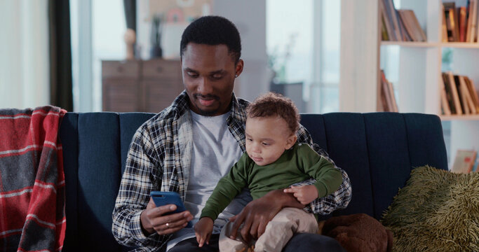 Daddy s lifestyle. Young african father using modern smartphone apps getting notifications holding baby son on hands. - Powered by Adobe