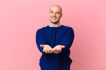 bald man smiling happily with friendly, confident, positive look, offering and showing an object or...