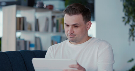 Young business casual guy staying home holding tablet lying on sofa videochatting on corporate meeting video conference indoors. Communications.