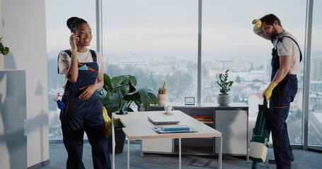 Lazy african young talkative woman dialing mobile phone while her tired male colleague janitor...