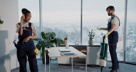 Lazy african young talkative woman dialing mobile phone while her tired male colleague janitor cleaning office alone using vacuum cleanner.