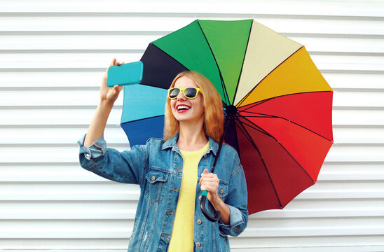 Portrait of happy smiling young woman taking a selfie by smartphone with colorful umbrella on a white background
