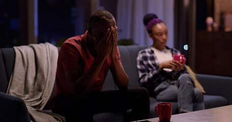 Couple in quarrel. Young serious african woman sitting with smartphone on couch. Her frustrated boyfriend crying in background learning divorce news.