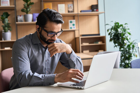 Latin Indian Serious Businessman Wearing Glasses And Headset Having Virtual Team Meeting Call, Remotely Working At Home Watching Online Learning Training Webinar In Remote Office.