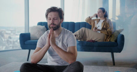 Caucasian couple relaxing at home, smoking cannabis weed and doing leisure things. Calm man doing meditation practice session while his girlfriend talking mobile phone.