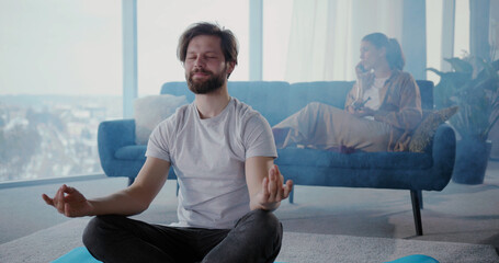Caucasian couple relaxing at home, smoking cannabis weed and doing leisure things. Calm man doing meditation practice session while his girlfriend talking mobile phone.