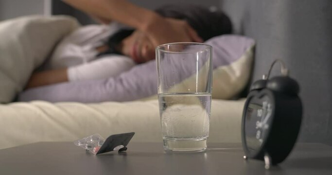 Young woman can't sleep at night because of insomnia or headache. Reaching out for pills and glass of water from bedside table. Health care concept.