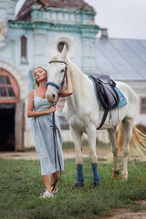 Beautiful woman in a blue dress hugs a white horse. Best friends. Photoshoot with a horse. Hobby love for animals. Country life. Romantic elegant girl tenderness. Saddle and bridle.