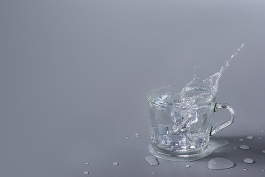 Crystal Clear Water With Ice In Transparent Glass Cup On A Gray Background. Splash And Splash