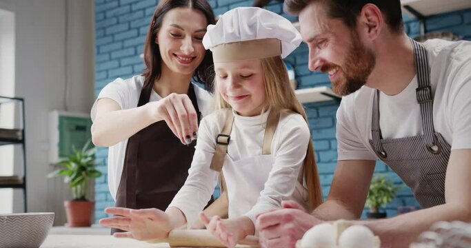 Happy family cooking making pizza together. Loving young parents helping cute little gir daughter to roll the dough, teaching kid to bake. Mom sprinkling flour on pastry.