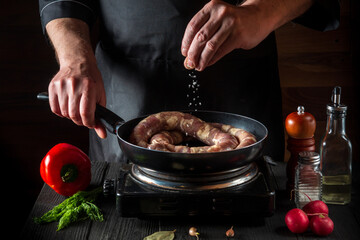 The chef adds salt to pan with raw meat sausage. Preparation for cooking sausages in the kitchen of a restaurant or cafe on table with vegetables and spices
