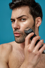 Obraz premium Closeup of brunette handsome guy looking at camera while using electric razor as a part of his morning routine isolated over blue background
