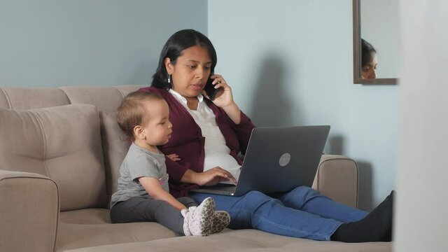 A Hispanic woman dressed in red and white is sitting in a couch working or studying online at a laptop and talking on phone together with a baby girl 