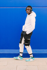 Young afro American black man wearing a white sweatshirt and headphones walking near a blue wall looking at camera. Modern look with confident attitude