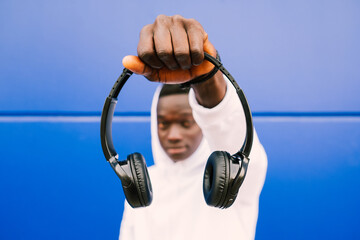 Young black man holding wireless headphones while wearing a white sweatshirt, against a blue wall...