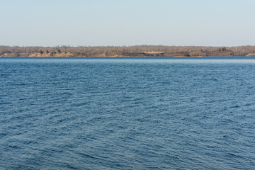 View of a blue lake, clear blue sky and opposite hilly shore with trees on a sunny windy spring day. Nature landscape background
