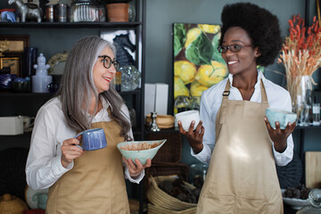 Smiling multiracial women in eyewear and apron holding decor and looking at each other. Two female sellers working together at modern store.