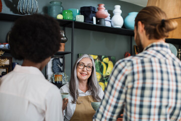 Positive grey-haired saleswoman consulting young multiracial couple at decor store. Back view of man and woman choosing products to buy.