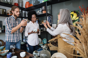 Happy multiracial customers bying original ceramic and glass products at modern decor shop. Competent mature saleswoman in beige apron showing various goods at store for young multiracial couple.