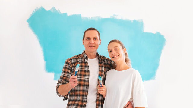 Happy couple doing repairs and painting walls in blue. Family standing with paint brushes, during apartment renovation concept.