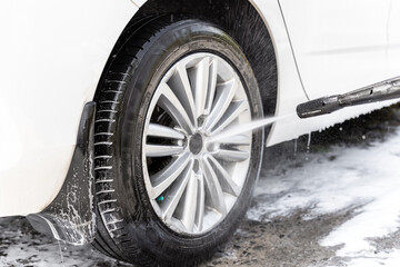 Close-up detail car wash with high pressure water equipment pump at home backyard outdoor on bright shiny summer day. Vehicle covered with foam shampoo chemical detergents during carwash self service