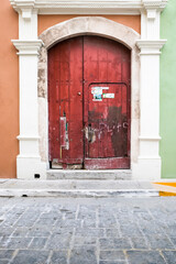 Close up of weathered large, tall, arched, red, double wooden doors with a smaller door cut into one side,  white columns, orange and light green walls of the Spanish colonial house, Campeche, Mexico.