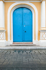 Fototapeta na wymiar Close up of very large, tall arched bright blue wooden double doors with a smaller door cut into one set in a yellow Spanish colonial house with decorative white columns, Campeche, Mexico