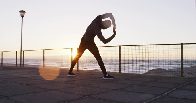 Focused african american man exercising outdoors, stretching by seaside at sunset
