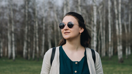 Close up of a woman dressed in linen, wearing sun glasses and a backpack, looking towards the cloudy sky. Concept: outdoor leisure, park walks