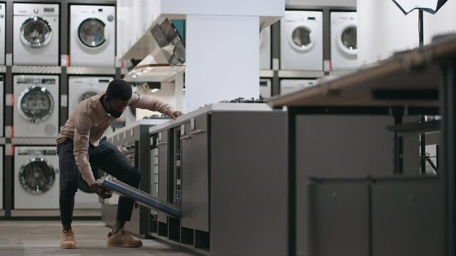 Black Guy Is Viewing Dishwasher In Trading Area Of Modern Home Appliance Store, Opening Door And Examining Inside