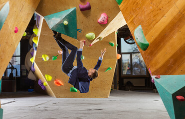 Young man in suit climbing difficult route on artificial wall in bouldering gym