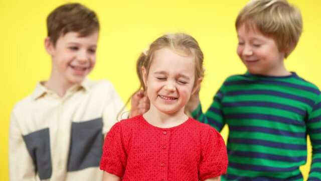 Portrait of cute little girl looking at camera as boys pulling pigtails at yellow background. Charming Caucasian sister suffering brothers mocking. Bullying and misbehave