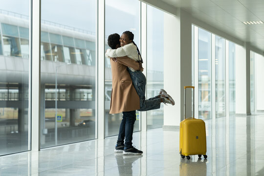 Happy Couple Meeting At Airport: Black Man Hugging Woman After Arrival In Terminal. Afro American Friends Or Lovers After Separation Due To Covid-19 Lockdown. Cheerful Female Fly To Loving Boyfriend