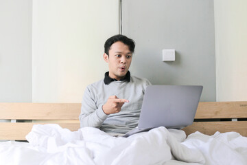 Surprised and shocked Asian man is working with his laptop on his cozy bed. Concept of successful freelancer lifestyle.