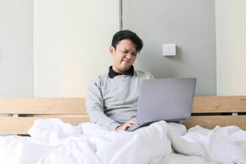 Young asian Man with a laptop in bed is confused and stress. Work from home concept