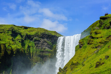 Tourists overlooking Iceland's Skogafoss waterfall surrounded by blue sky and greenery