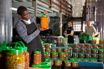 Focused African-American man working in food producing factory, inspecting and arranging glass jar with pickled olives