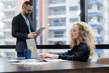Business woman sitting on the chair and using laptop to see information from internet in the modern high level office building. Background is a man standing beside windows. Teamwork brainstorm concept