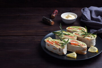 Rolls of thin pita bread and red salted salmon with lettuce leaves on a black ceramic plate, dark wooden background. Copy space.