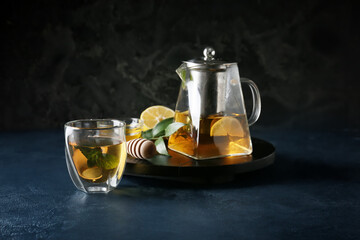 Teapot and glass of tasty tea with lemon and mint on dark background