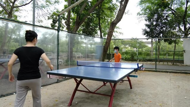 Asian 40s cute mother and her son playing table tennis outdoor  in the park , the boy wearing  facemask to prevent coronavirus covid-19 during pandemic . Child and mother concept.