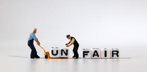 Two miniature men moving the cube of 'UN' word from cube with 'FAIR' word to cart. White cube with words and miniature people.
