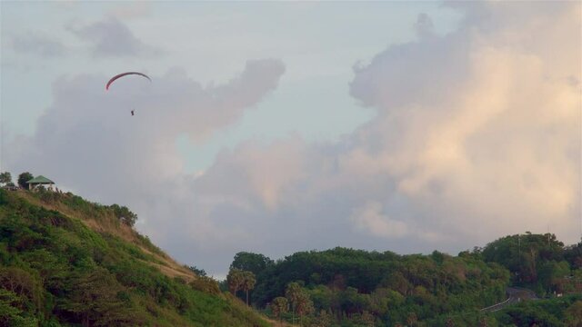 The paraglider soars by parachute on the green mountains. In the distance there is an observation deck with people watching the sunset.