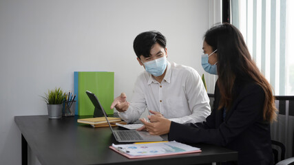 Two young business people in protective mask discussing project and sharing ideas in office room.